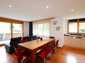 Una cocina moderna con encimera de madera y electrodomésticos integrados, un comedor con mesa de madera y sillas rojas, y un gran ventanal con cortinas.
