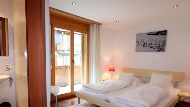 A bedroom with a bed, a nightstand, a window with curtains, a wall-mounted picture, and a wooden door. - Aramis Appartement/Fewo 6-Bettwohnung Nr. 2 by Interhome (Saas-Fee)
