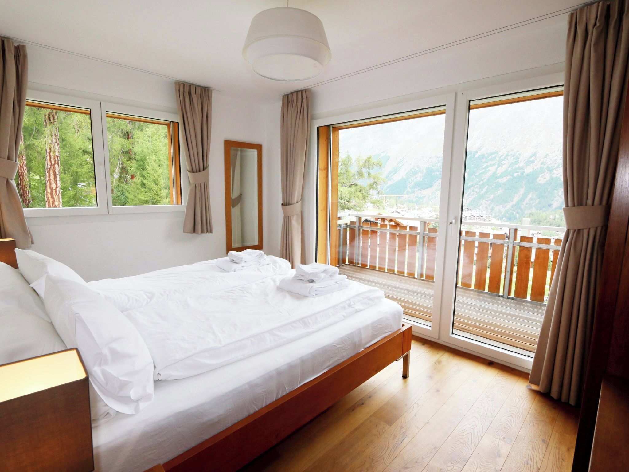 A hotel room with a large bed, a wooden headboard, a large window with a view of mountains, and a balcony with a wooden railing.