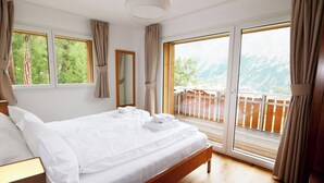 Quarto de hotel com uma cama grande, cabeceira de madeira, uma janela ampla com vista para montanhas e uma varanda com grade de madeira.