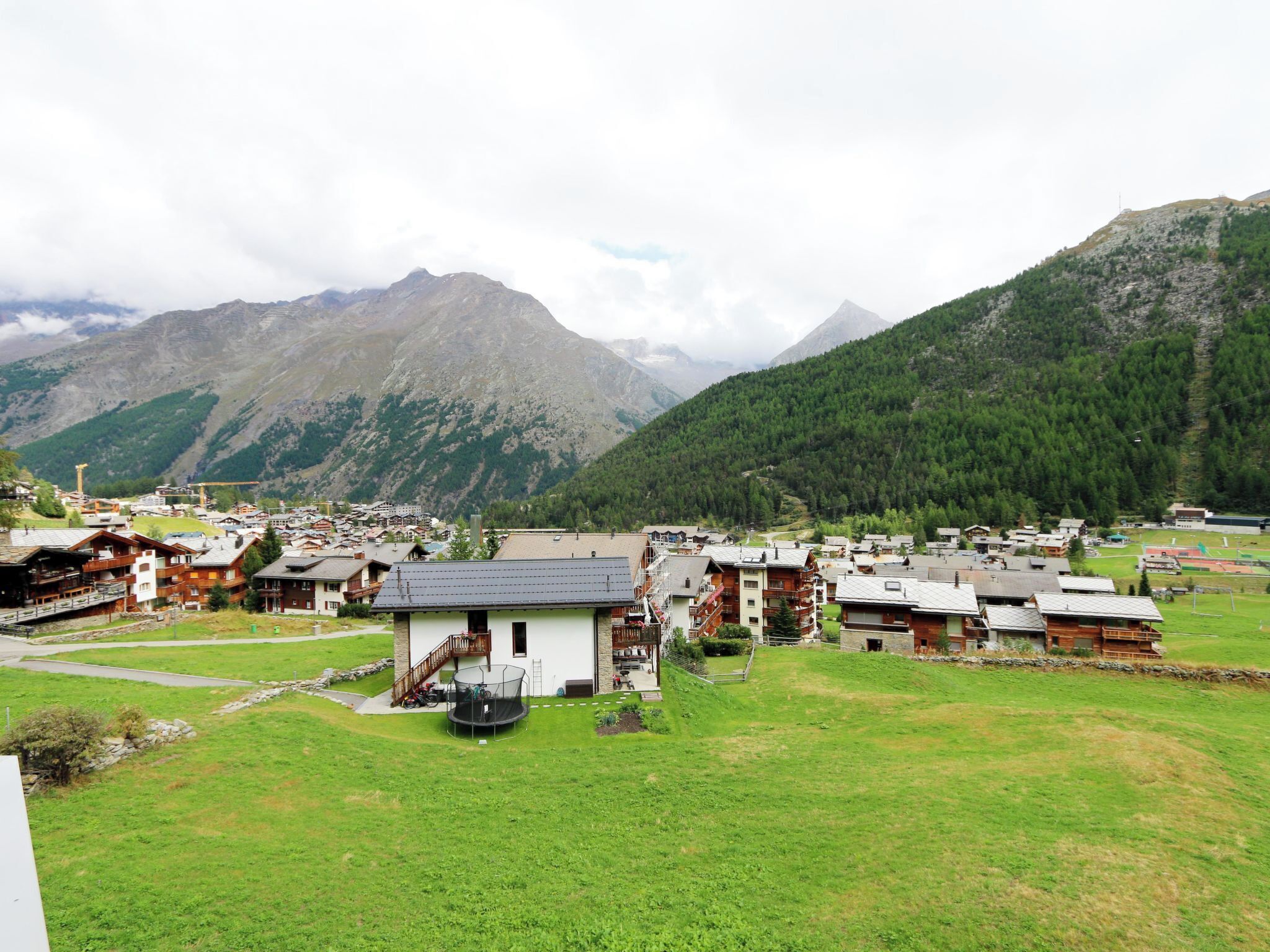 Un village de montagne aux maisons traditionnelles en bois, avec un bâtiment central blanc doté d’un balcon, et offrant une vue dégagée sur les sommets environnants.