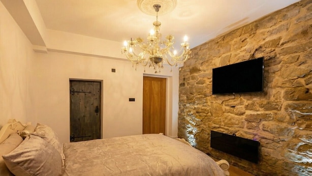 A bedroom with a large bed, a chandelier, a stone wall, a wooden door, and a mounted TV. - Stone-Walled 1-Bed with Fireplace · Sleeps 2 by St Edward`s Church, Castle Donington (Castle Donington)