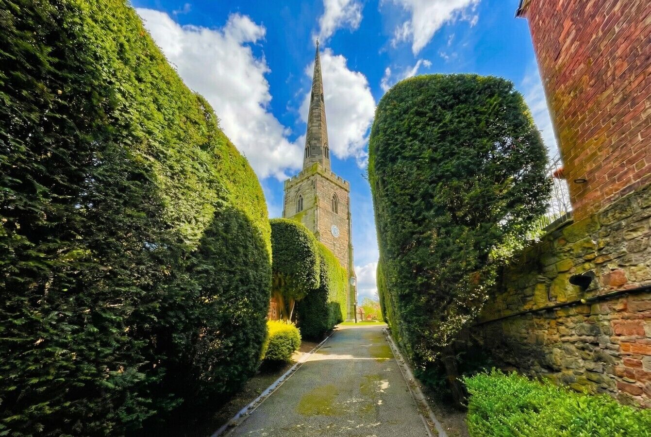 Ein schmaler Pfad, gesäumt von ordentlich geschnittenen Hecken, der zu einer Kirche mit einem hohen Spitzturm führt.