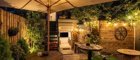 Un espace extérieur aménagé en terrasse, avec des tables et des chaises en bois, un grand parasol et une enseigne indiquant « BAR TRICTOR'S ».