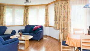 A living room with a blue sofa, a wooden coffee table, and a dining area with a table and chairs. - Amaryllis Appartement/Fewo 4-Bettwohnung A by Interhome (Saas-Grund)