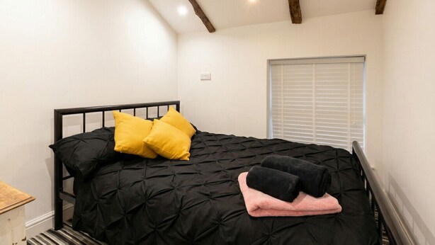 Une chambre à coucher avec un lit noir, deux oreillers jaunes, une couverture noire et rose, et une fenêtre avec des stores.