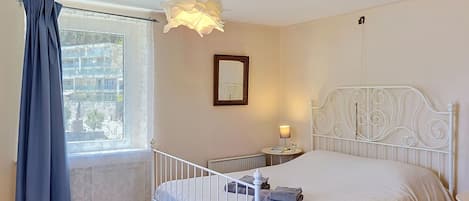 A bedroom with a white metal bed frame, a bedside table with a lamp, a window with curtains, and a radiator.