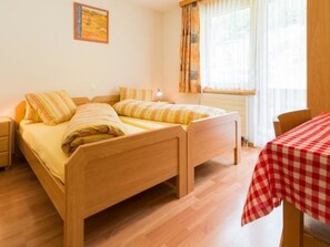 Ein Zimmer mit zwei Holzbetten, einem kleinen Tisch mit kariertem Tischdecken und einem Fenster mit Vorhängen.