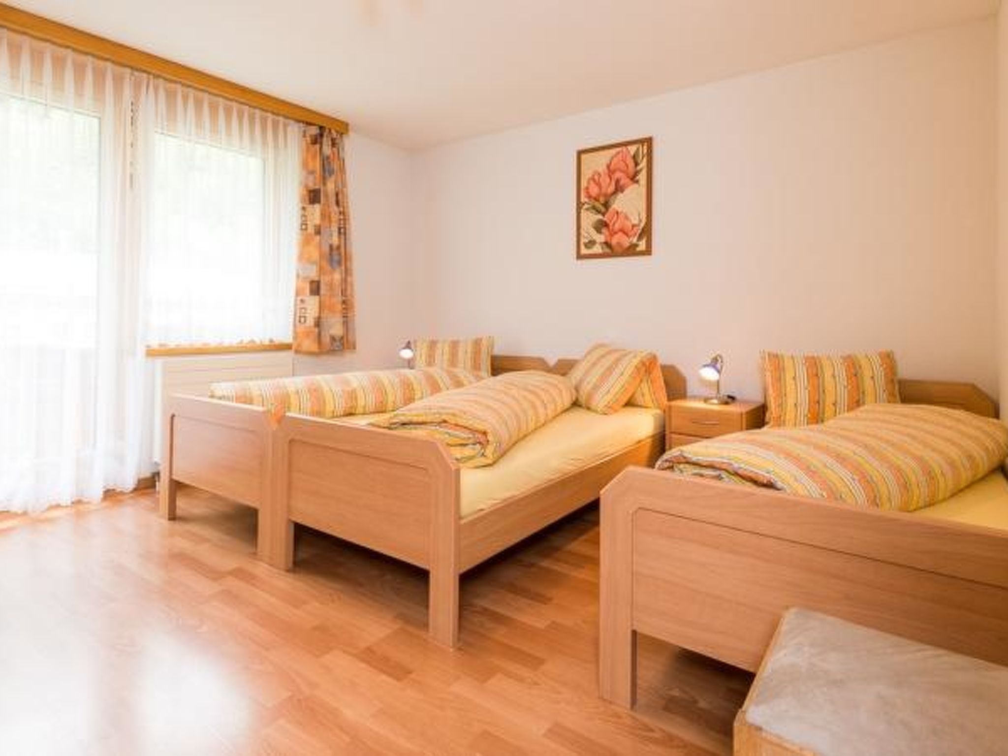 A room with three wooden beds, a large window with sheer curtains, and a single wall-mounted lamp.