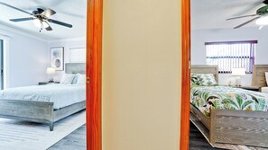 Quarto com duas camas, um ventilador de teto, janela com persianas e mesa de cabeceira com abajur.