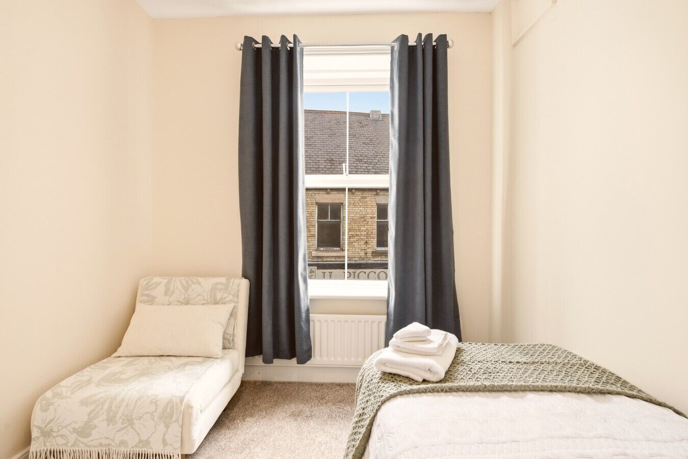 Ein Schlafzimmer mit zwei Betten, einem fensterbelaubten Fenster, einem Heizkörper und Blick auf ein Gebäude.