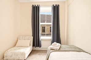 A bedroom with two beds, a window with curtains, a radiator, and a view of a building outside.
