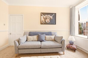 A living room with a grey sofa, patterned cushions, a framed animal painting, a small table with a lamp, and a window with a view.