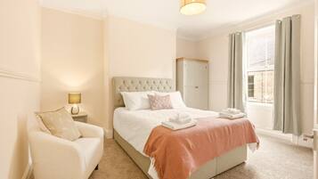 A bedroom with a bed, a chair, a nightstand, a lamp, a wardrobe, and a window with curtains.