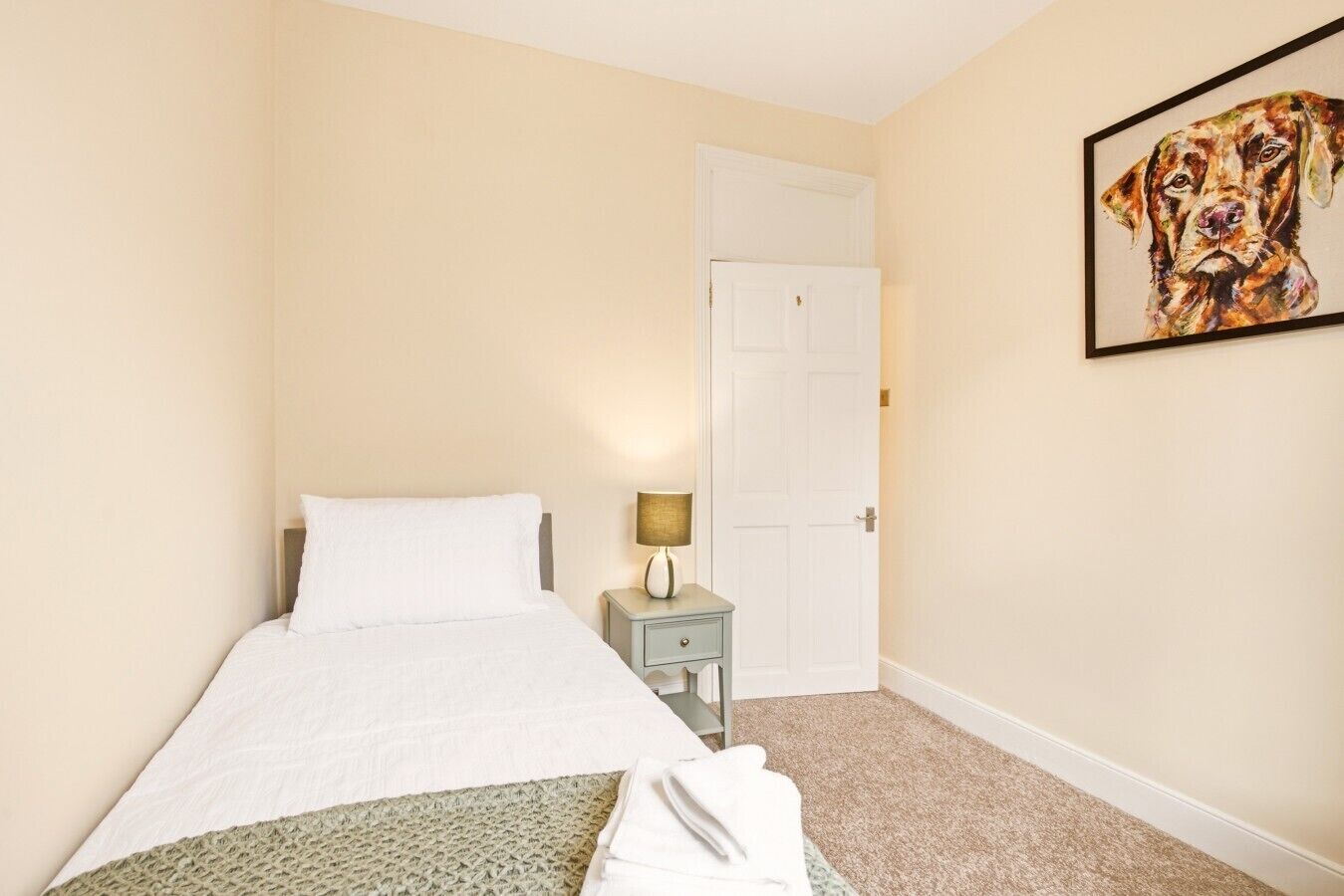 A hotel room with a bed, a nightstand with a lamp, a door, and a framed picture on the wall.