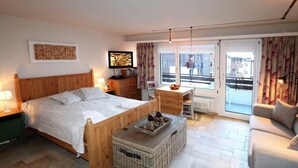 A bedroom with a wooden bed, a small table, a wicker basket, a sofa, and a view of the outside through a large window. - Sarazena Appartement/Fewo 2-Bettwohnung O by Interhome (Saas-Fee)