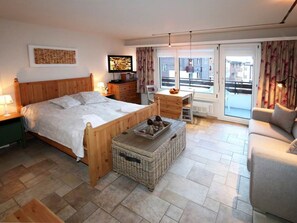 Ein Schlafzimmer mit einem hölzernen Bett, einem kleinen Tisch, einem Korbwannenstuhl, einer Couch und Blick ins Freie durch ein großes Fenster.