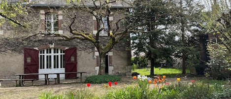 A building with a red door and windows, surrounded by a garden with various plants and flowers.