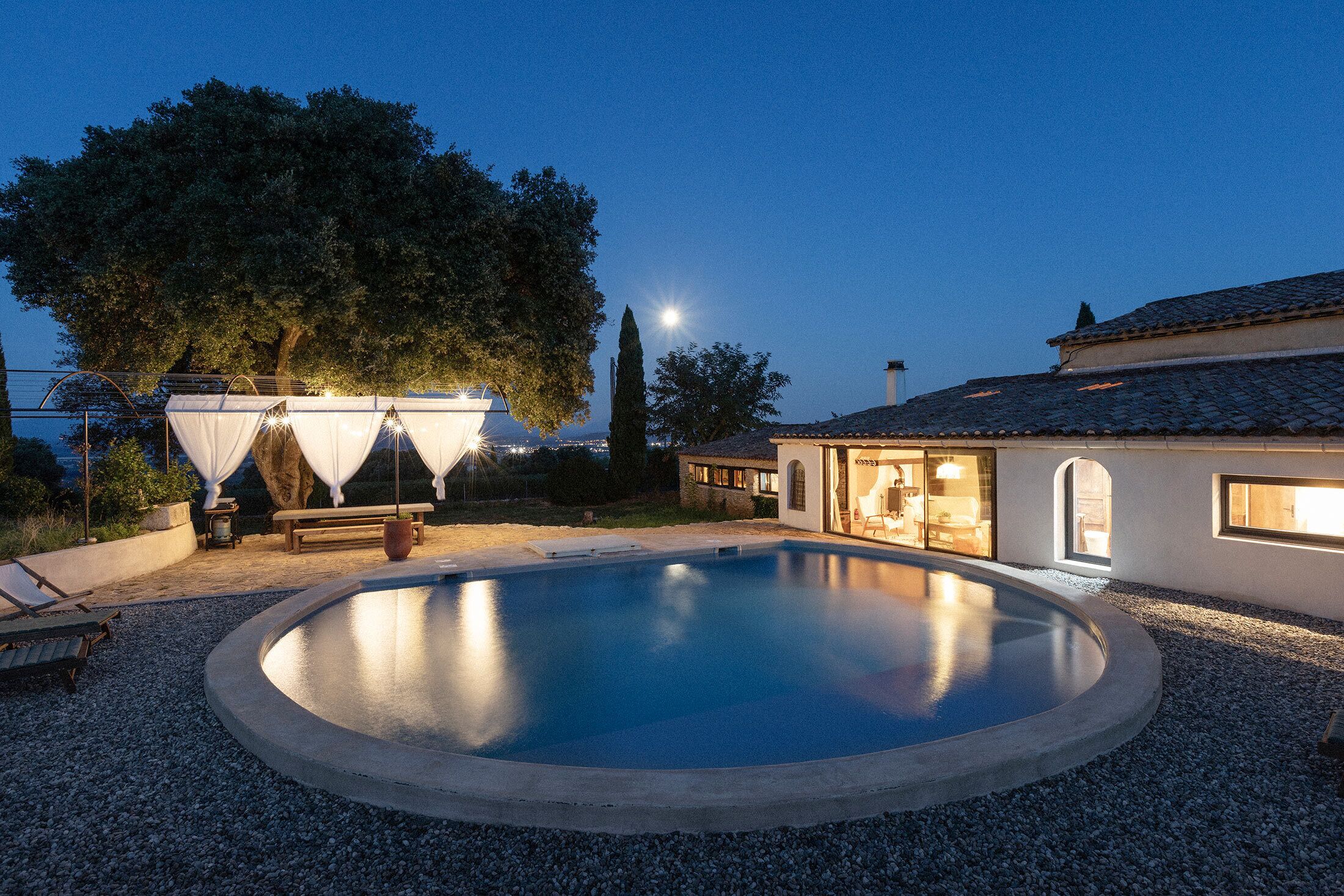 A circular swimming pool with a surrounding gravel area, a bench, and a covered seating area with string lights.