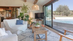 A living room with a sofa, a coffee table, and a TV. There are plants and a large window with a view of a pool and trees. - La Sardagne | Charming House in the Heart of the Garrigue, Near Vallon-Pont-d'Arc (Bourg-Saint-Andéol)