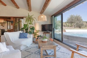 A living room with a sofa, a coffee table, and a TV. There are plants and a large window with a view of a pool and trees. - La Sardagne | Charming House in the Heart of the Garrigue, Near Vallon-Pont-d'Arc (Bourg-Saint-Andéol)