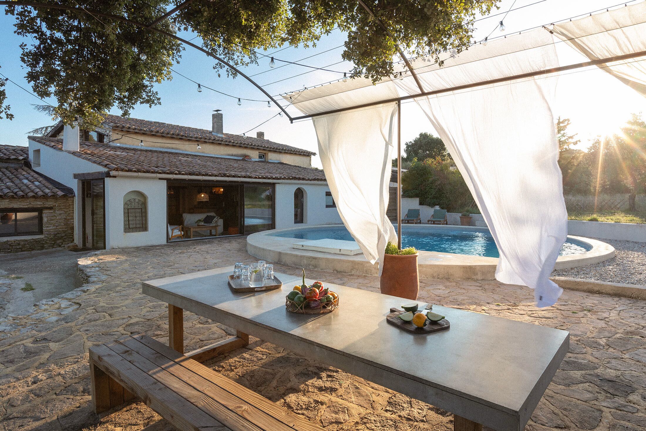 Un espace extérieur aménagé pour les repas, avec une table dressée pour deux personnes, une piscine et, au fond, un bâtiment doté d’un toit recouvert de tuiles.