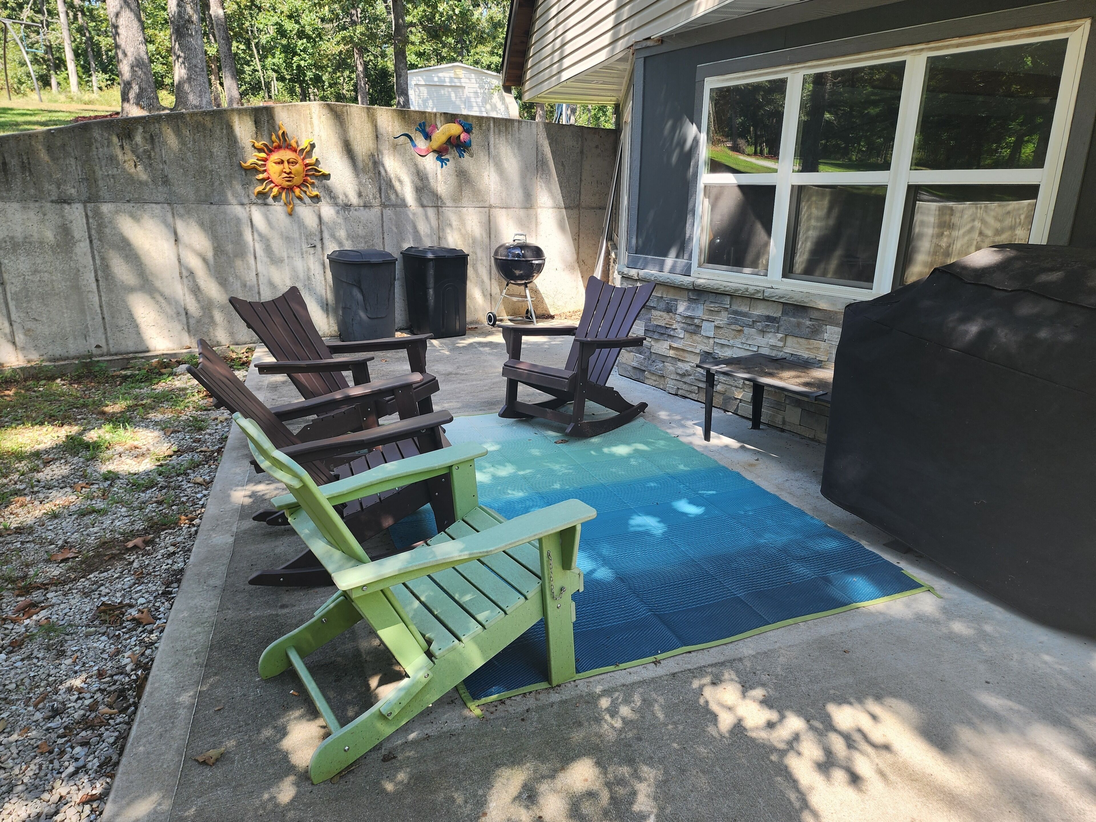 Een terras buiten met een blauwe mat, twee schommelstoelen, een tafel en een barbecue.