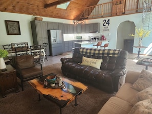 A spacious living area with a leather sofa, a coffee table, and a dining set. A kitchen is visible in the background with modern appliances.