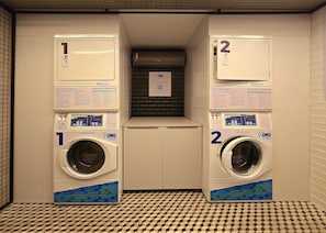 A laundry room with two washing machines, numbered 1 and 2, and a small shelf with a QR code. - Get By Side By Hosppedar (João Pessoa)