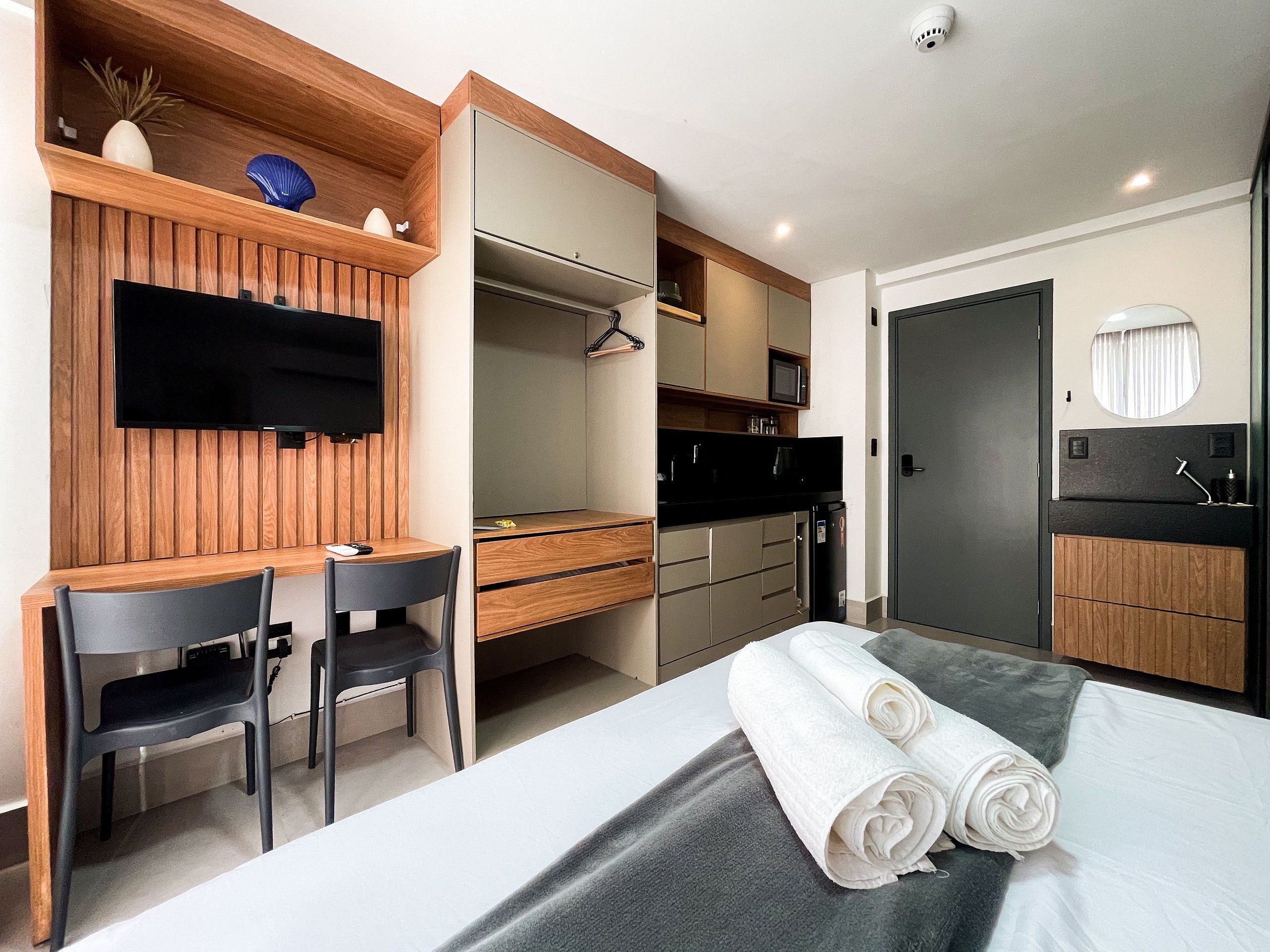 Une chambre d’hôtel moderne avec un bureau en bois, un téléviseur à écran plat, un lit recouvert d’une couverture grise et une salle de bain équipée d’un miroir et d’un lavabo.