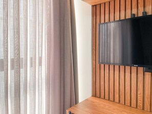 A modern room with a flat-screen TV mounted on a wooden panel wall, a window with sheer curtains, and a wooden floor. - Get By Side By Hosppedar (João Pessoa)