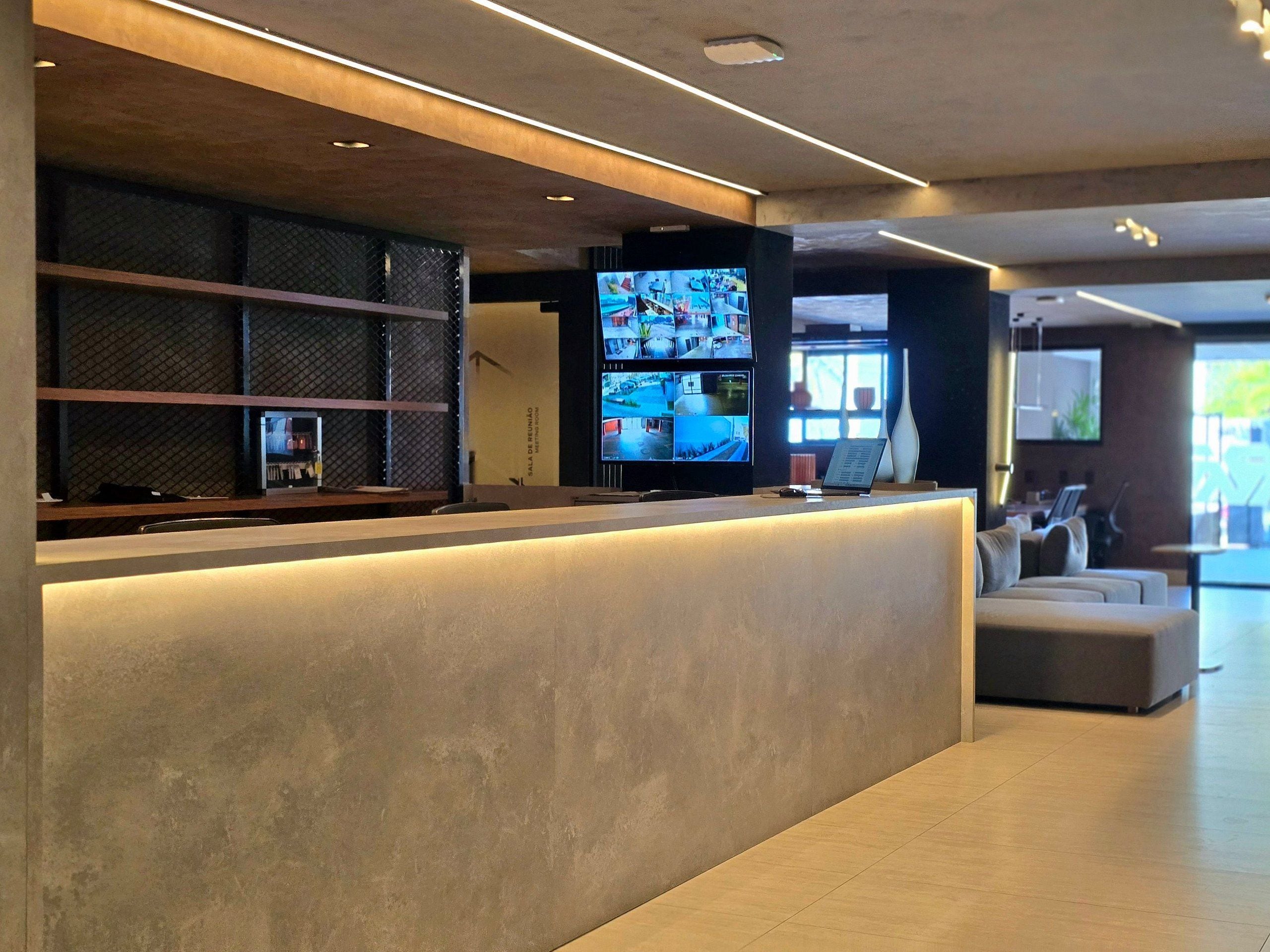 Zona de recepción con diseño moderno, que incluye un mostrador de recepción, un televisor de pantalla plana grande mostrando múltiples cámaras y un área de estar con sofá y sillones.