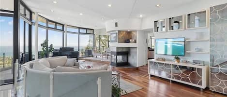 A modern living room with a curved sofa, a fireplace, a television, and a large window overlooking a scenic view.