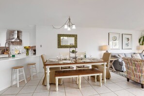 A dining area with a wooden table, white chairs, and a bench. There is a kitchen area with a stove and sink to the left. - BEACHFRONT! Beautiful pool, hot tub, tennis, pickle &LOTS of restaurants nearby! (St. Augustine)