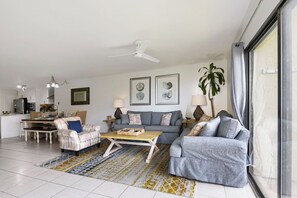 A modern living room with a dining area, a ceiling fan, and a large window. - BEACHFRONT! Beautiful pool, hot tub, tennis, pickle &LOTS of restaurants nearby! (St. Augustine)