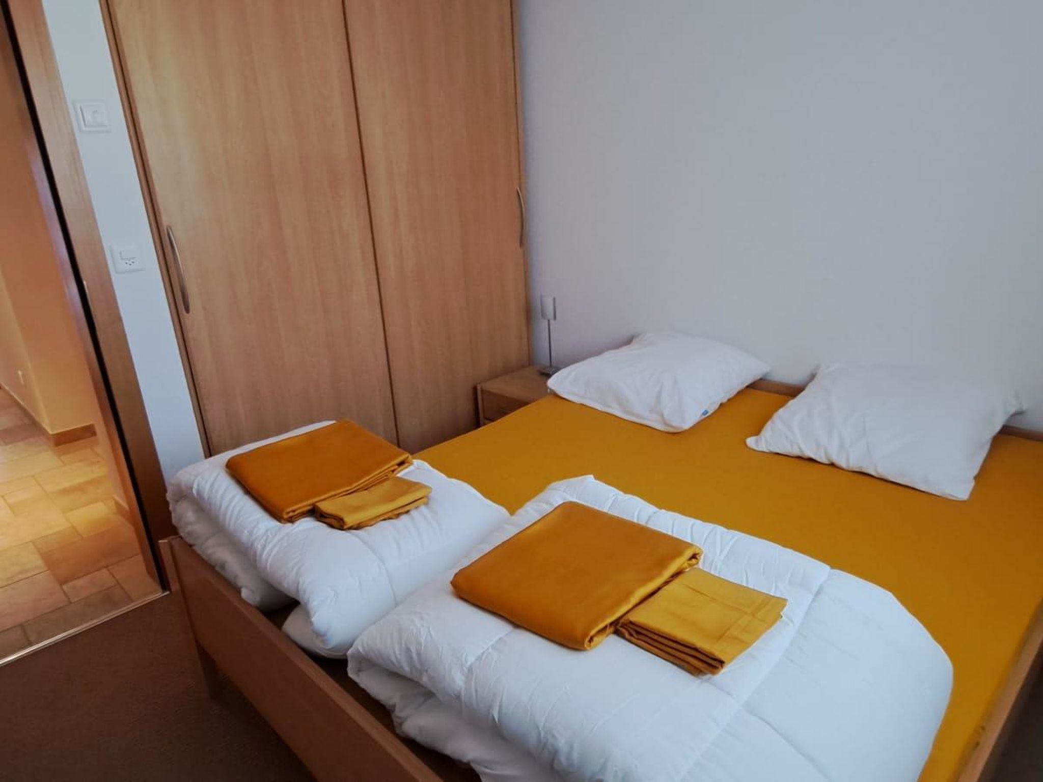 Une chambre avec deux lits, chacun recouvert d’une literie blanche et d’une couverture jaune. On y trouve une armoire en bois et une table de chevet avec une lampe.