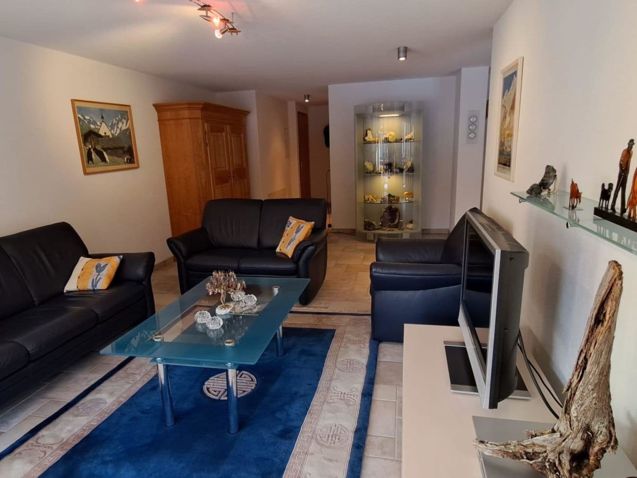 A living room with two black sofas, a glass coffee table, a TV, and a decorative shelf with figurines.