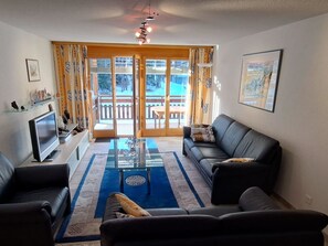 A living room with a sofa, a coffee table, a TV, and a view of a snowy outdoor area through glass doors. - Grossus Moos A22 Appartement/Fewo Appartement/Fewo by Interhome (Saas-Fee)