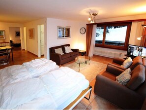 Quarto de hotel com uma cama, dois sofás, uma mesa pequena, um relógio e uma janela com cortinas.