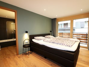 Ein modernes Schlafzimmer mit einem großen Bett, einem kleinen Sessel, einem Spiegel und Blick ins Freie.