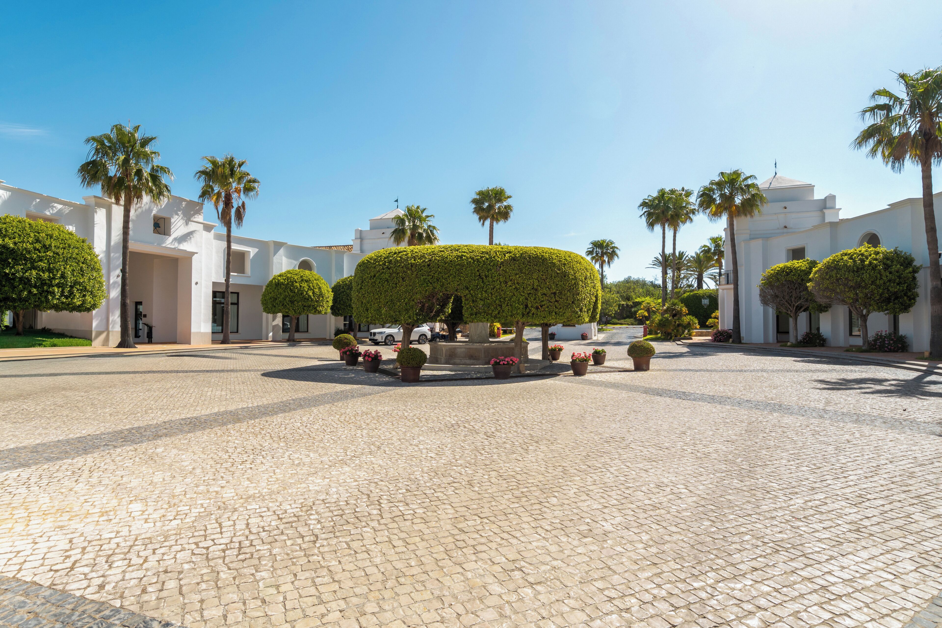 Un patio con una fuente central, rodeado de edificios blancos y palmeras.