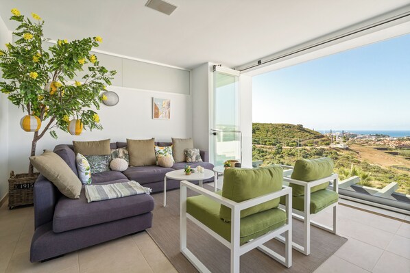 Ein modernes Wohnzimmer mit Sofa, Sesseln, Couchtisch und einem großen Fenster mit Blick auf eine hügelige Landschaft.