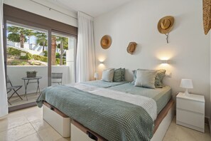 Ein Schlafzimmer mit Bett, Nachttischen, einem Balkon mit Stuhl und Blick auf Palmen.