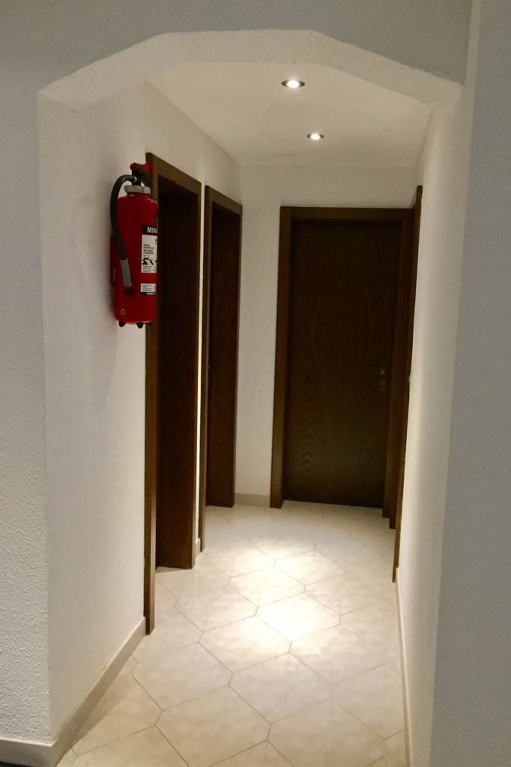 Un pasillo con extintor montado en la pared, puertas de madera y suelo de baldosas.