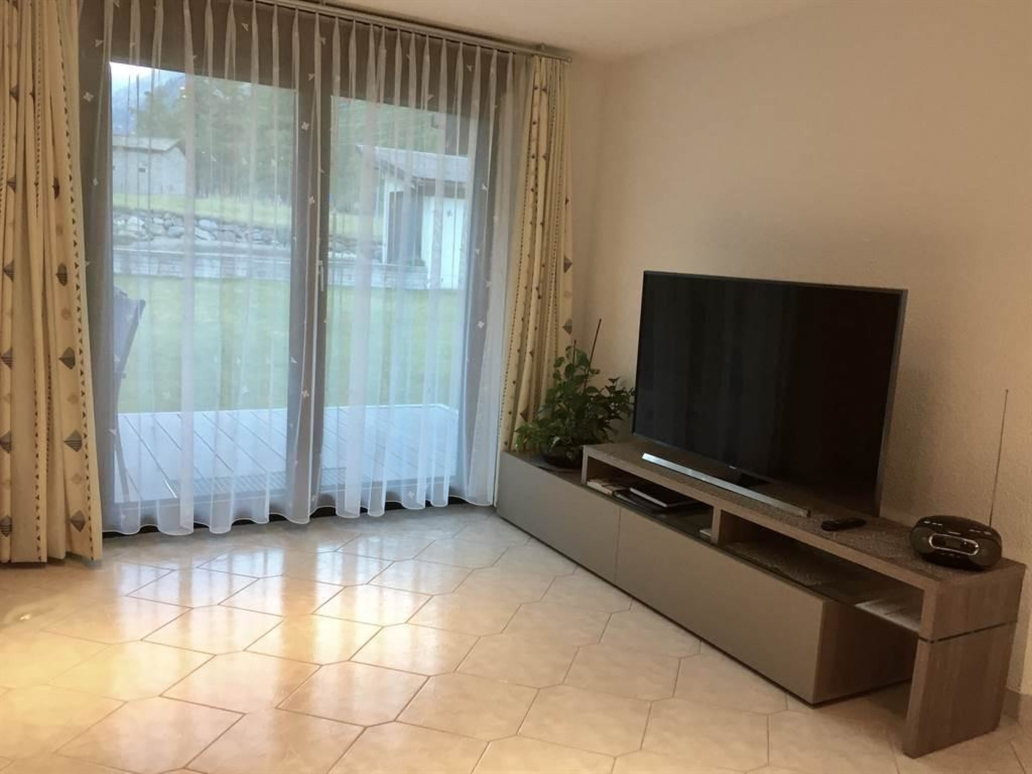 Un saló amb televisió de pantalla plana sobre un suport de fusta, una porta corredissa de vidre amb cortines translúcides i un sòl de rajola.