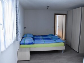 Ein Einzelbett mit Matratze und Kissen, ein kleiner Nachttisch, ein Kleiderschrank und ein Fenster mit Vorhängen.