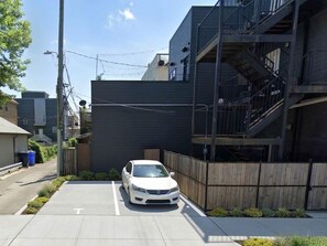 Un aparcament amb una furgoneta blanca, un edifici amb una escala de foc i un carrer amb una tanca i vegetació.