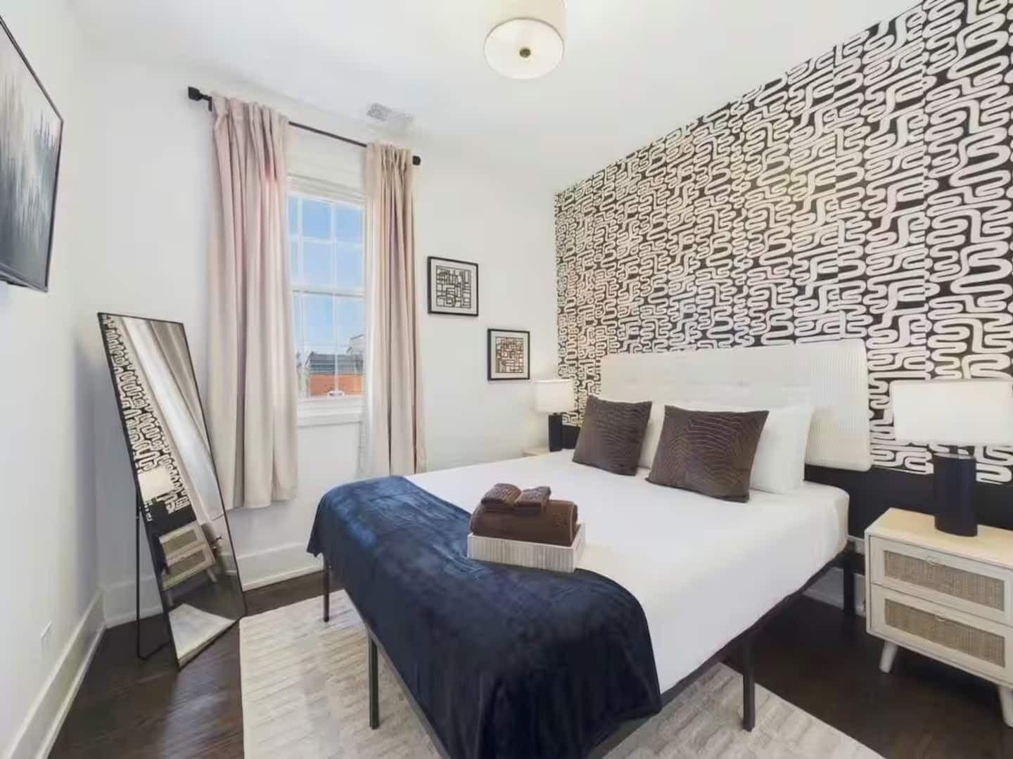 A hotel room with a bed, a bedside table, a mirror, a window with curtains, and a patterned wall.