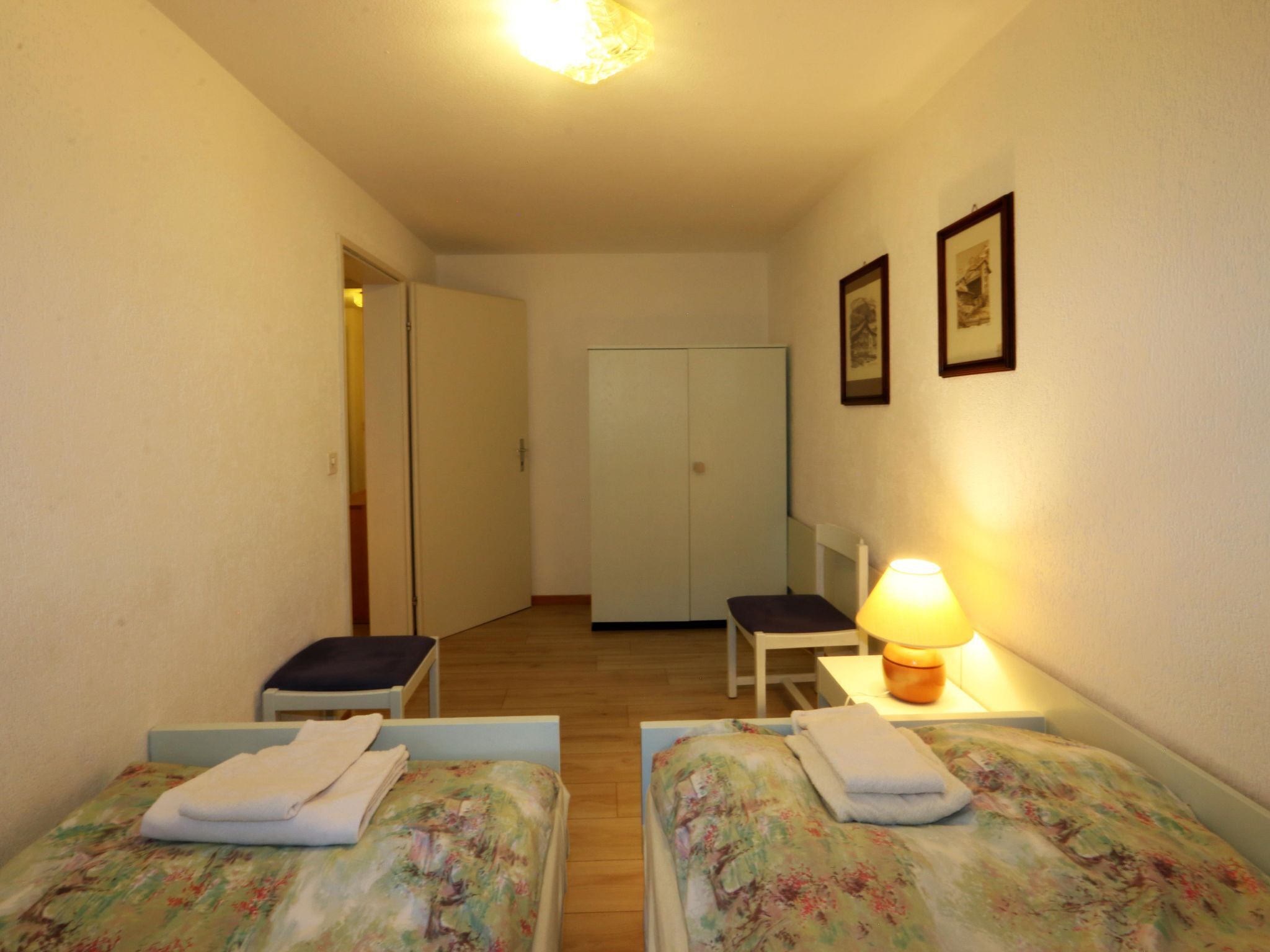 A small, well-lit room with two single beds, a chair, a desk with a lamp, and a wardrobe.