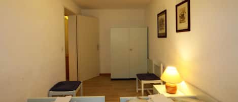 A small, well-lit room with two single beds, a chair, a desk with a lamp, and a wardrobe.